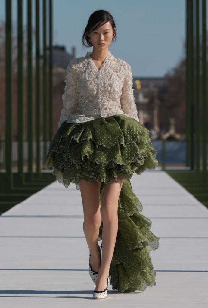Mujer desfilando un conjunto de falda verde larga y una blusa de mangas larga blanca, tacones blancos con polka dots